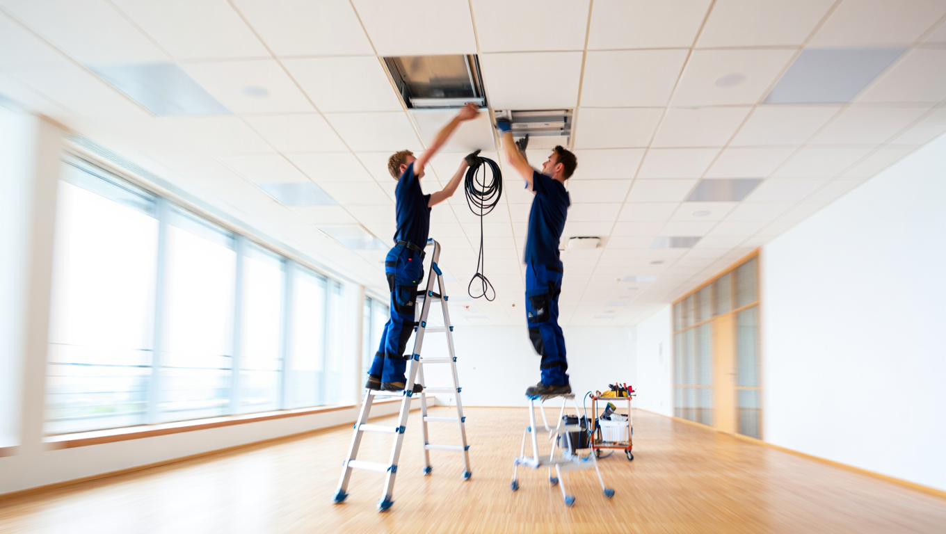 Zwei Handwerker installieren ein Deckenlicht in einem modernen Büro, während sie auf Leitern stehen. Das Bild zeigt eine helle, offene Raumgestaltung mit Holzfußboden.