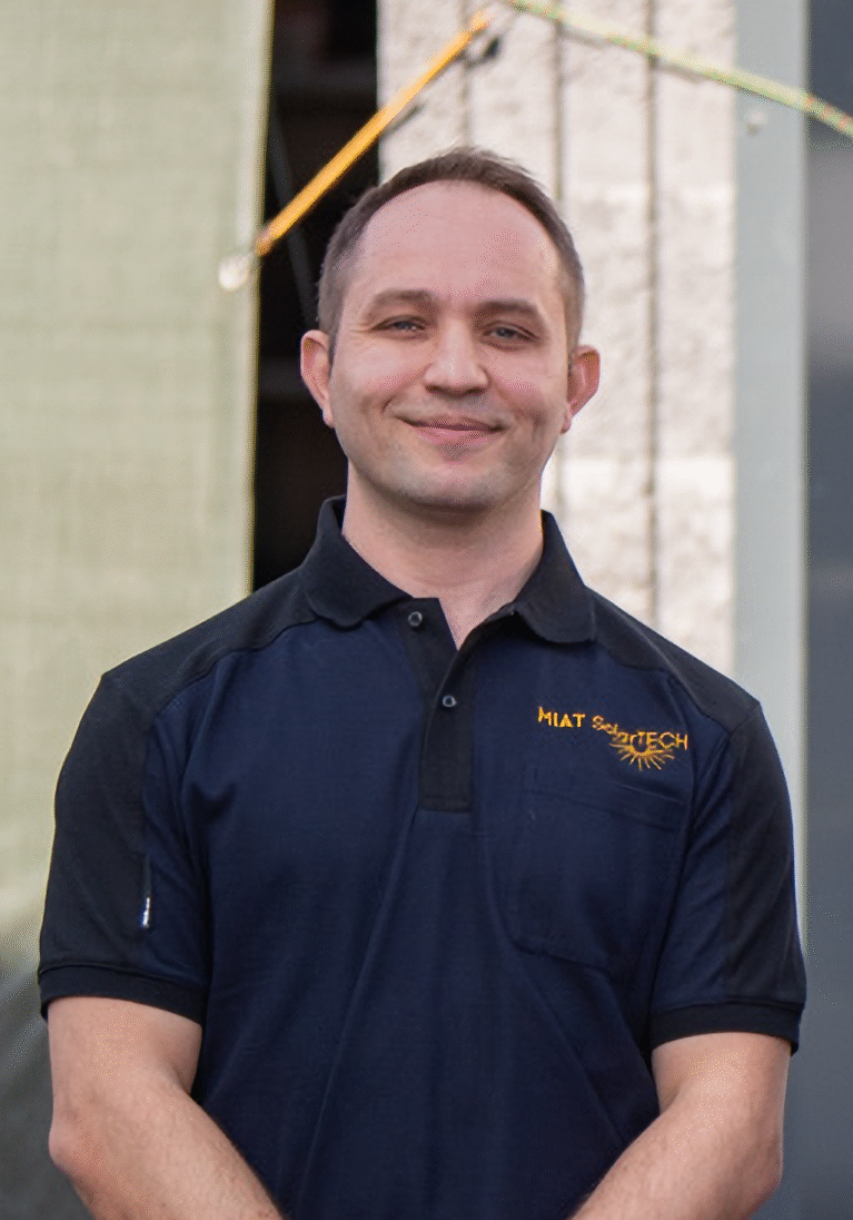 Mann in schwarzem Polo-Shirt mit Logo steht vor einem Gebäude und lächelt.