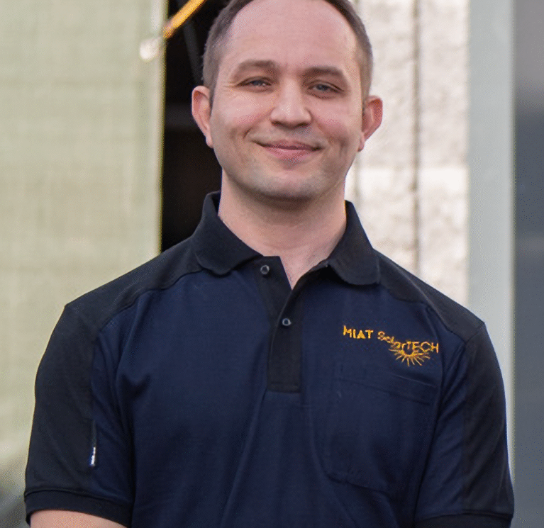 Mann in schwarzem Polo-Shirt mit Logo steht vor einem Gebäude und lächelt.