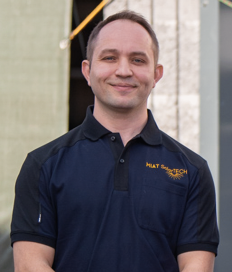 Mann in schwarzem Polo-Shirt mit Logo steht vor einem Gebäude und lächelt.