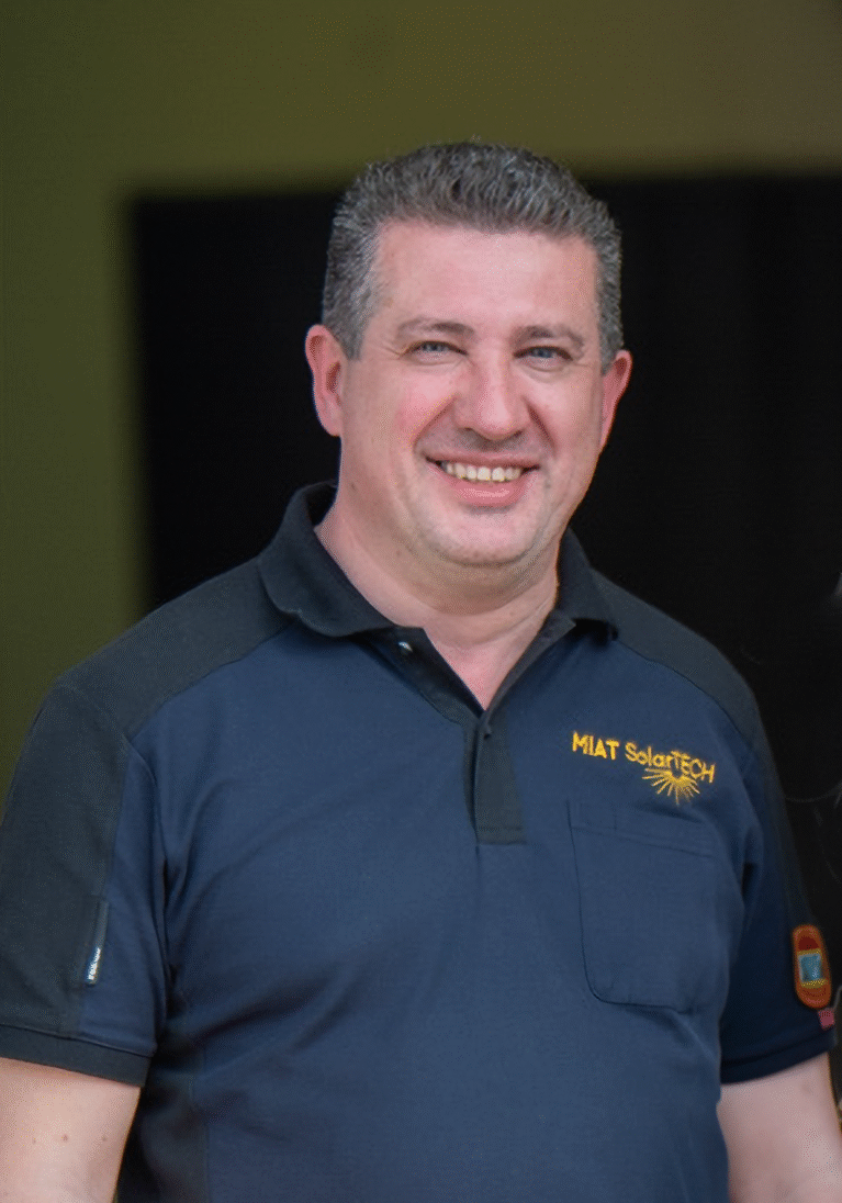 Lächelnder Mann in einem blauen Polo-Shirt mit Logo, steht vor einer dunklen Hintergrundwand.