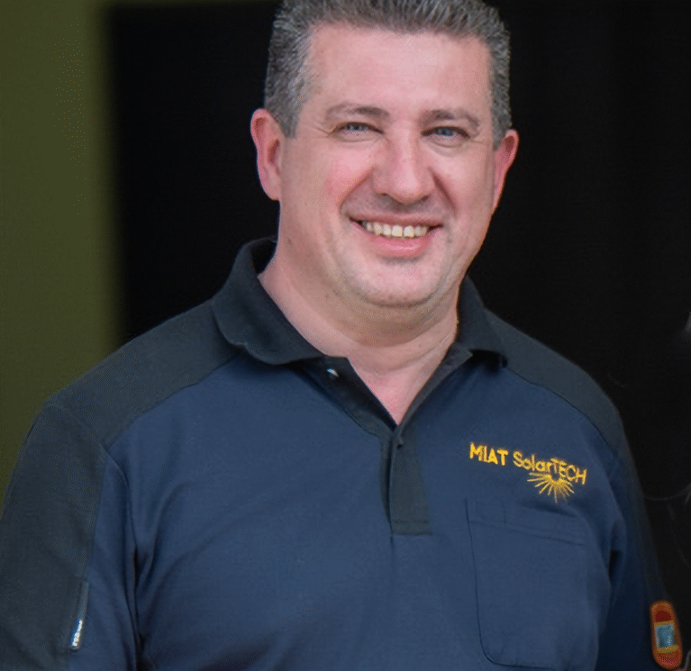 Lächelnder Mann in einem blauen Polo-Shirt mit Logo, steht vor einer dunklen Hintergrundwand.